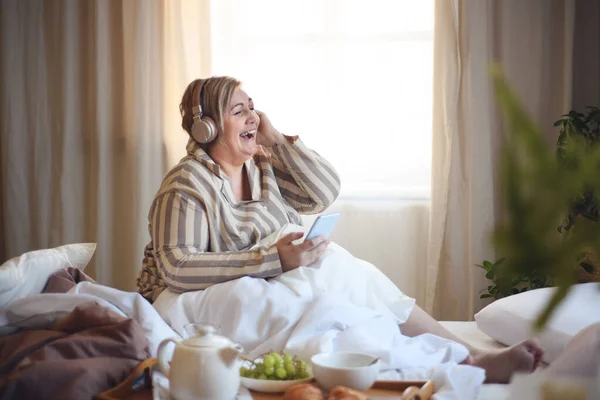 Glad överviktig kvinna med hörlurar och smartphone äter frukost i sängen hemma. — Stockfoto