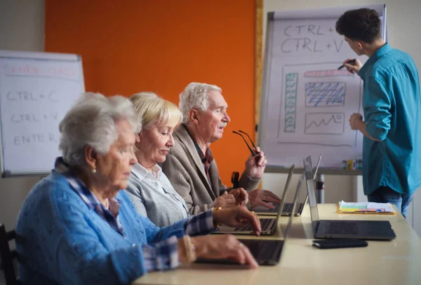 Grupo de personas mayores en un hogar de jubilados con un instructor joven aprendiendo juntos en clase de informática —  Fotos de Stock
