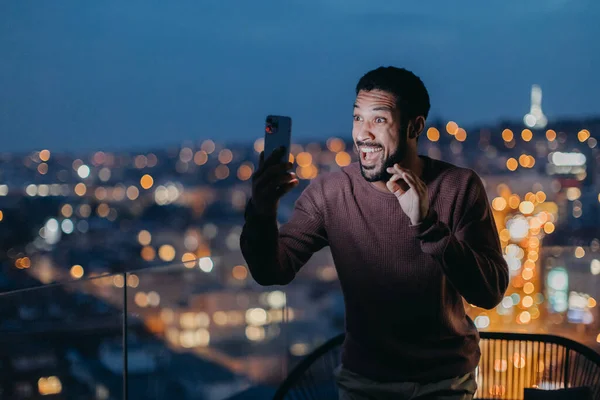 Wesoły młody człowiek robi selfie na balkonie z widokiem na miasto. — Zdjęcie stockowe