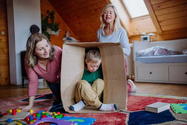 Down sendromlu bir çocuk. Annesi ve büyükannesi evde kutuyla oynuyorlar.. — Stok fotoğraf