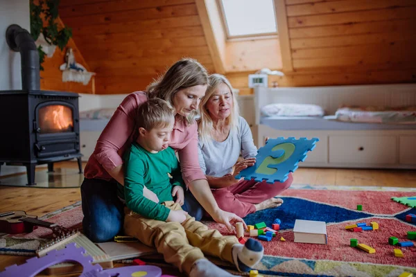 Gutt med Downs syndrom sitter på gulvet og leker med moren og bestemoren hjemme – stockfoto