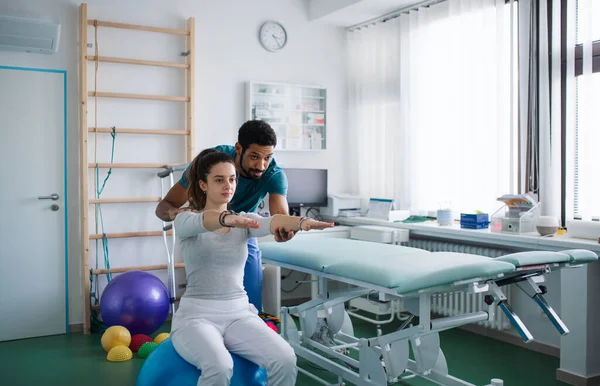 Joven fisioterapeuta masculino haciendo ejercicio con una paciente joven sobre el balón en una sala física —  Fotos de Stock
