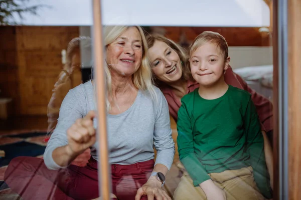 다운 증후군 이 있는 아이와 어머니와 할머니가 앉아서 집에서 창밖을 내다보고 있는 모습. — 스톡 사진