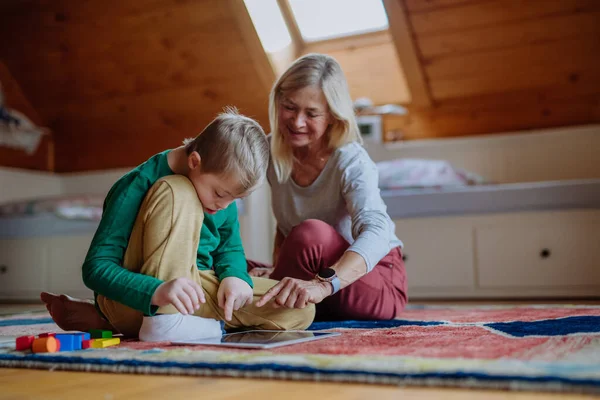 Kind met Down syndroom zitten op de vloer en het gebruik van tablet met oma thuis. — Stockfoto