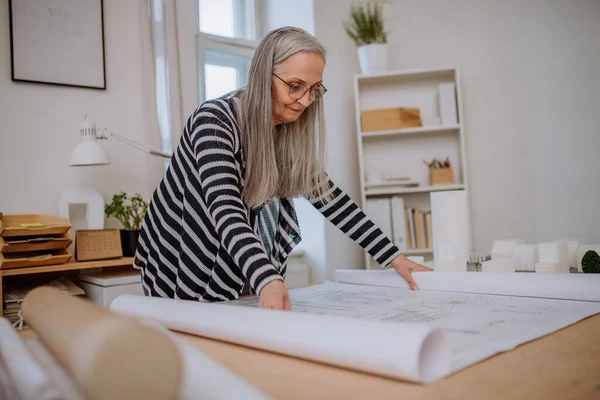 Senior woman architect with model of houses looking at blueprints in office. — Stock Photo, Image