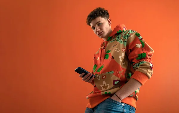 Estúdio de moda retrato de um jovem com capuz com smartphone posando sobre fundo laranja. — Fotografia de Stock