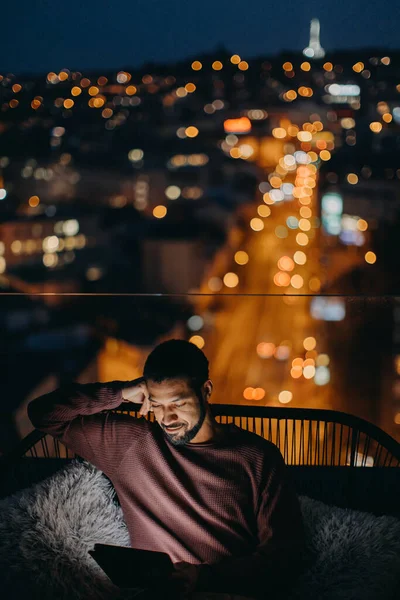 Молодой африканский американец сидит на балконе с видом на город и использует планшет ночью — стоковое фото