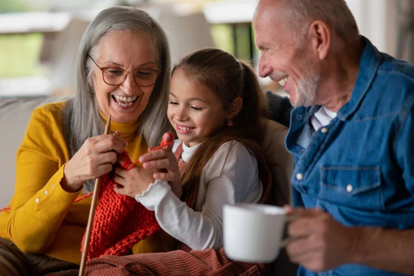 Niña sentada en el sofá con sus abuelos y aprendiendo a tejer en casa. — Foto de Stock