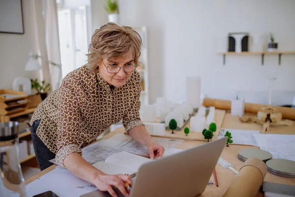 Старша жінка-архітектор з моделлю будинків, що працюють на ноутбуці в офісі . — стокове фото