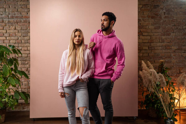 Fashion studio portrait of a happy young couple in hoodie posing over pink background.