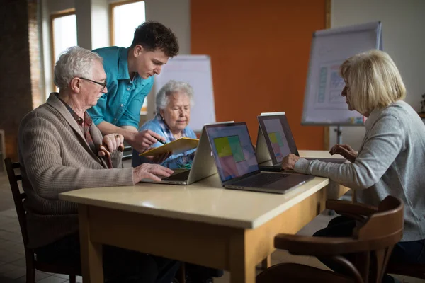 Grupo de personas mayores en un hogar de jubilados con un instructor joven aprendiendo juntos en clase de informática —  Fotos de Stock