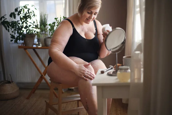 Kilolu bir kadın evde oturup bacaklarına krem sürüyor.. — Stok fotoğraf