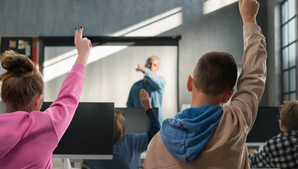 Rear view of school kids raising hands during computer calss at school. — ストック写真