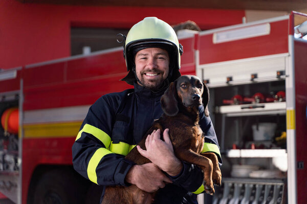 Happy firefighter man holding dog and looking at camera with fire truck in background