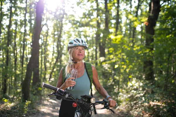 Happy senior kobieta rowerzysta pitnej wody z butelki na świeżym powietrzu w lesie. — Zdjęcie stockowe