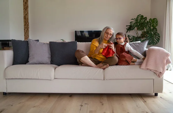 Liten flicka som sitter på soffan med sin mormor och lär sig att sticka inomhus. — Stockfoto
