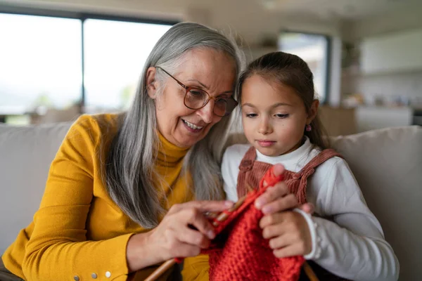 Menina sentada no sofá com a avó e aprendendo a tricotar dentro de casa. — Fotografia de Stock