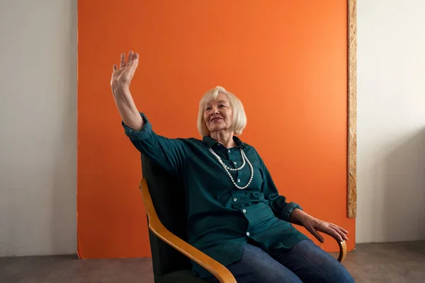 Smiling elegant elderly woman sitting in armchair and waving to someone. — Stock Photo, Image