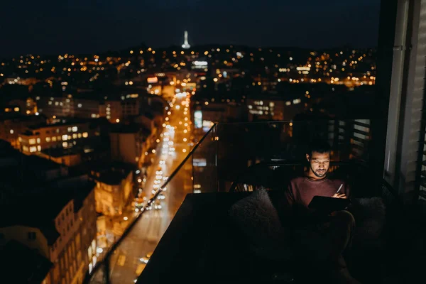 Молодой африканский американец сидит на балконе с видом на город и использует планшет ночью — стоковое фото