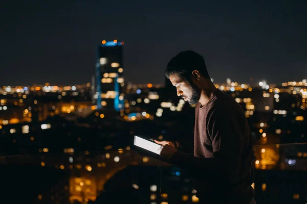 Młody Afroamerykanin stojący na balkonie z widokiem na miasto i korzystający w nocy z tabletu — Zdjęcie stockowe