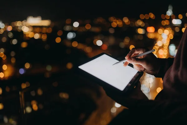 Nahaufnahme eines Mannes, der nachts auf einem Balkon mit Blick auf die Stadt steht und ein Tablet benutzt — Stockfoto