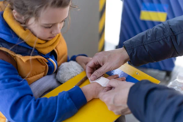 Voluntário ajudando a criança refugiada ucraniana na estação de trem, dando gesso à mão ferida. — Fotografia de Stock