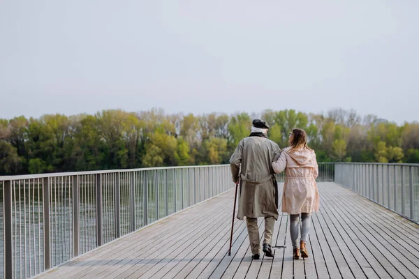Rear view of senior man with daughter outdoors on a walk on pier by river. — Foto de Stock