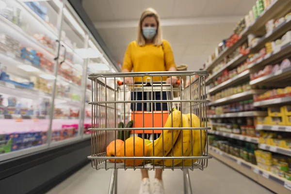 Jonge vrouw in het gezicht masker met trolley winkelen in supermarkt, inflatie concept. — Stockfoto