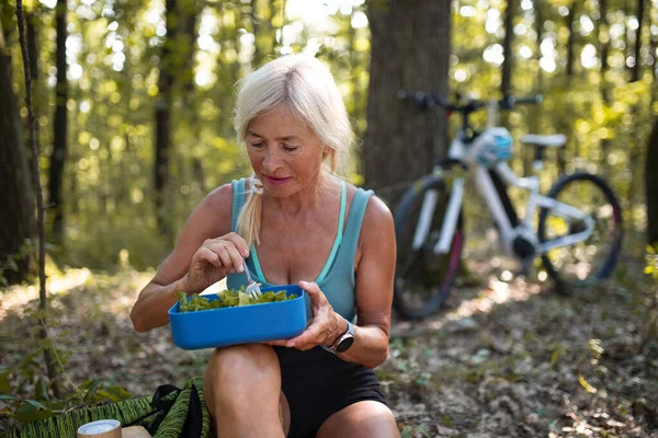 Χαρούμενη ηλικιωμένη γυναίκα ποδηλάτης αναπαύεται, κάθεται και τρώει σνακ σε εξωτερικούς χώρους στο δάσος. — Φωτογραφία Αρχείου