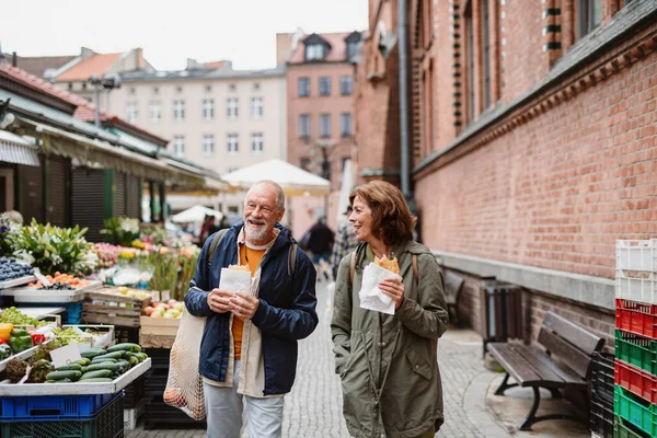 Happy senior couple tourists with snack in town on outdoor market. — Fotografia de Stock