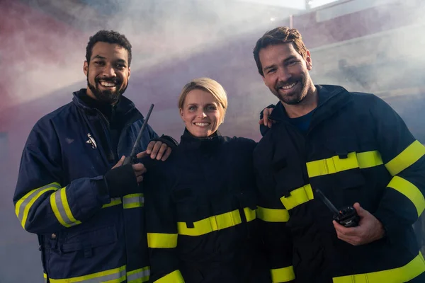 Happy firefighters crew with fire station and truck in background looking at camera.