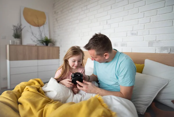 Padre poniendo el despertador con su hija pequeña con síndrome de Down en la cama en casa. — Foto de Stock