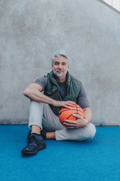 Mature man with baskatball sitting on courtoutdoors and looking at camera. — Foto Stock