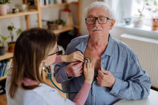 Médico visitando a un hombre mayor y examinándolo con estetoscopio en casa, escuchando latidos del corazón. — Foto de Stock