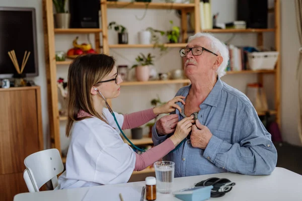 Médico visitando a un hombre mayor y examinándolo con estetoscopio en casa, escuchando latidos del corazón. — Foto de Stock