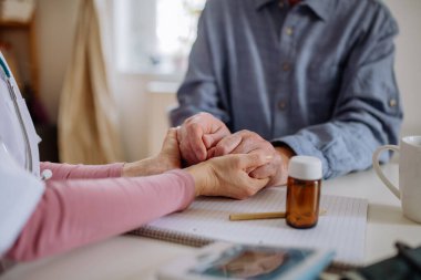 Close-up of doctor holding hands of senior patient and consoling him during medical visit at home.