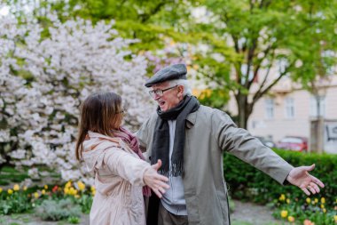 Adult daughter with outstretched hands meeting her senior father outdoors in park on spring day.