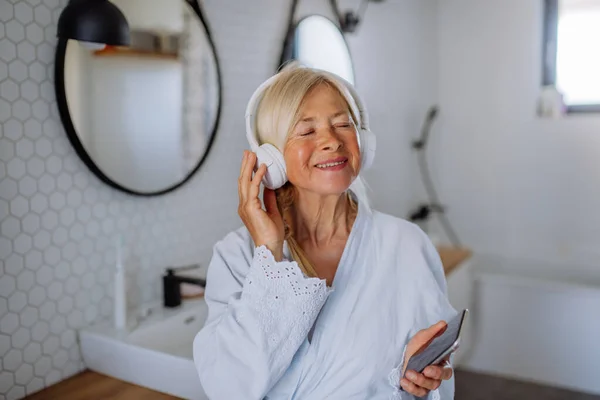 Schöne Seniorin im Bademantel hört Musik im Bad, Entspannungs- und Wellness-Konzept. — Stockfoto
