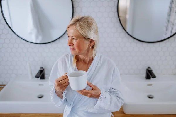 Beautiful senior woman in bathrobe drinking tea in bathroom, relax and wellness concept. — Foto de Stock