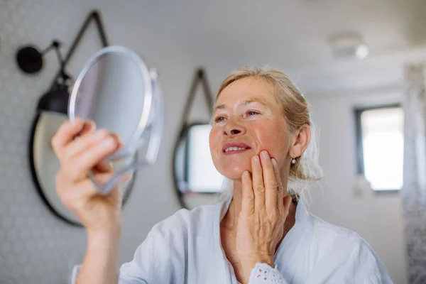 Bella donna anziana in accappatoio guardando specchio e applicando crema viso naturale in bagno, concetto di cura della pelle. — Foto Stock