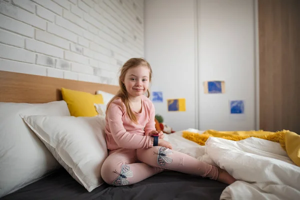 Niña feliz con síndrome de Down mirando a la cámara y sentada en la cama en casa. —  Fotos de Stock