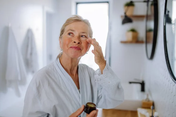 Schöne Seniorin im Bademantel Anwendung natürlicher Gesichtscreme im Bad, Hautpflege und morgendliches Routinekonzept. — Stockfoto