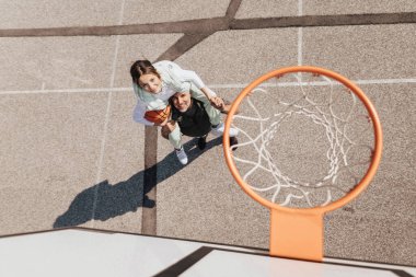 Father and teenage daughter playing basketball outside at court, high angle view above hoop net.