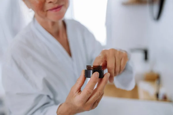 Mujer mayor en albornoz aplicando crema facial natural en baño, cuidado de la piel y concepto de rutina matutina. — Foto de Stock