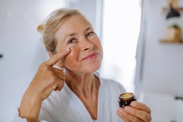 Belle femme âgée en peignoir appliquant crème naturelle pour le visage dans la salle de bain, soins de la peau et concept de routine du matin. — Photo