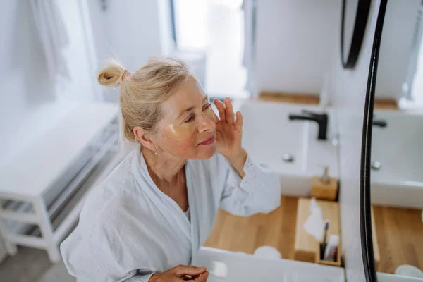 Beautiful senior woman in bathrobe, applying eye patches for puffiness while looking in the mirror — Stok Foto