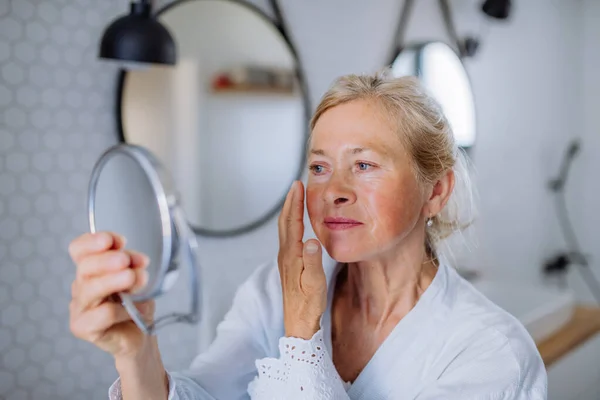 Beautiful senior woman in bathrobe looking at mirror and applying natural face cream in bathroom, skin care concept. — Stok Foto