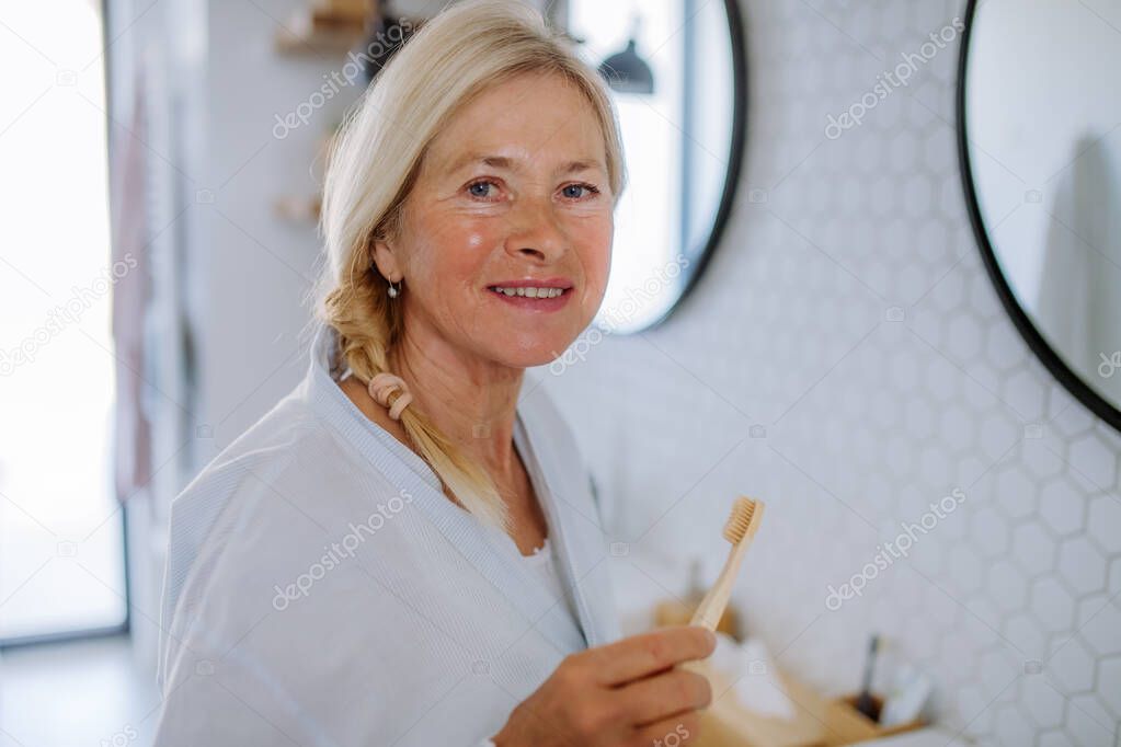 Beautiful senior woman in bathrobe brushing teeth with eco wooden toothbrush inbathroom, sustainable lifestyle.