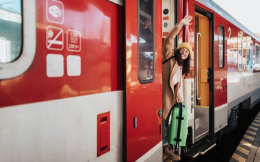 Tren istasyonunda tren kapısında durup el sallayan, bavullu mutlu genç bir gezgin kadın.
