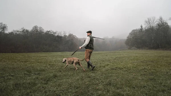 Hunter man met hond in traditionele schietkleren op het veld met shotgun. — Stockfoto